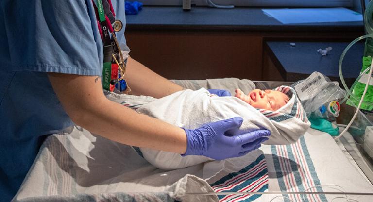 Newborn wrapped in a blanket is gently lifted by a gloved health professional in the neonatal intensive care unit