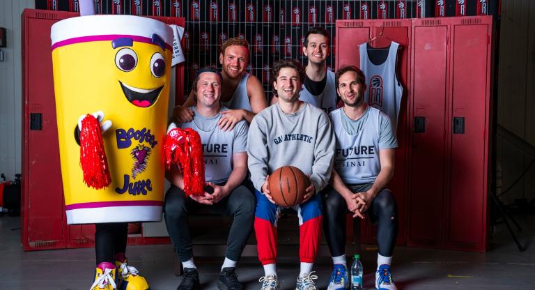 Five basketball teammates arranged in two rows in front of red lockers, smiling next to the Booster Juice mascot. Three sit in the front with the middle player holding a basketball, and two stand behind