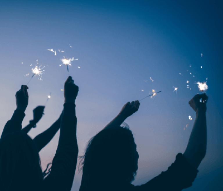 Silhouettes of three people holding sparklers against a twilight sky.