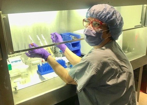a woman working in a laboratory-like setting, wearing scrubs and a mask.