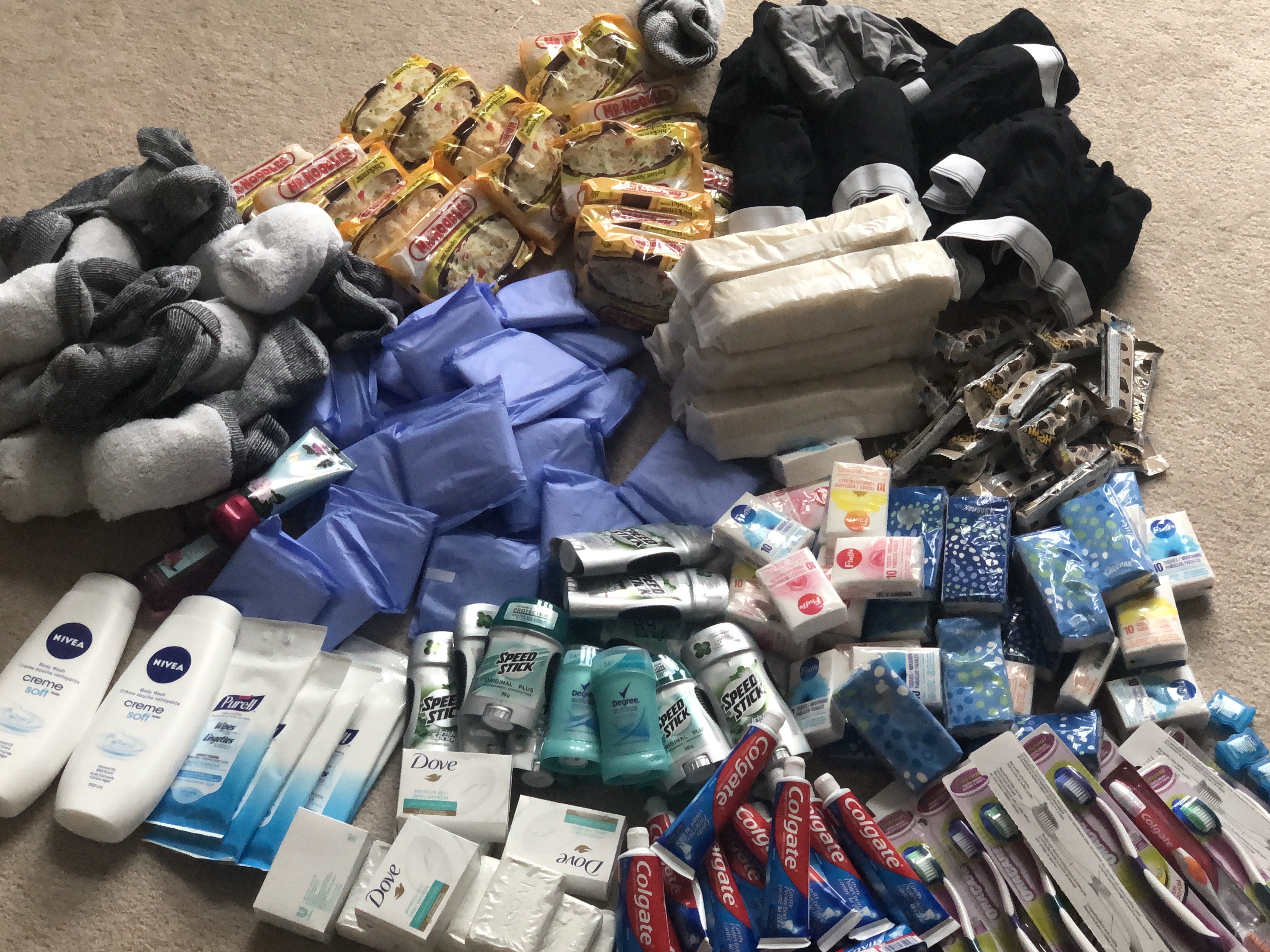 A pile of several dozen donated personal products, including soap, toothbrushes and socks along with packaged noodles and crackers and other snacks on a carpeted floor. 