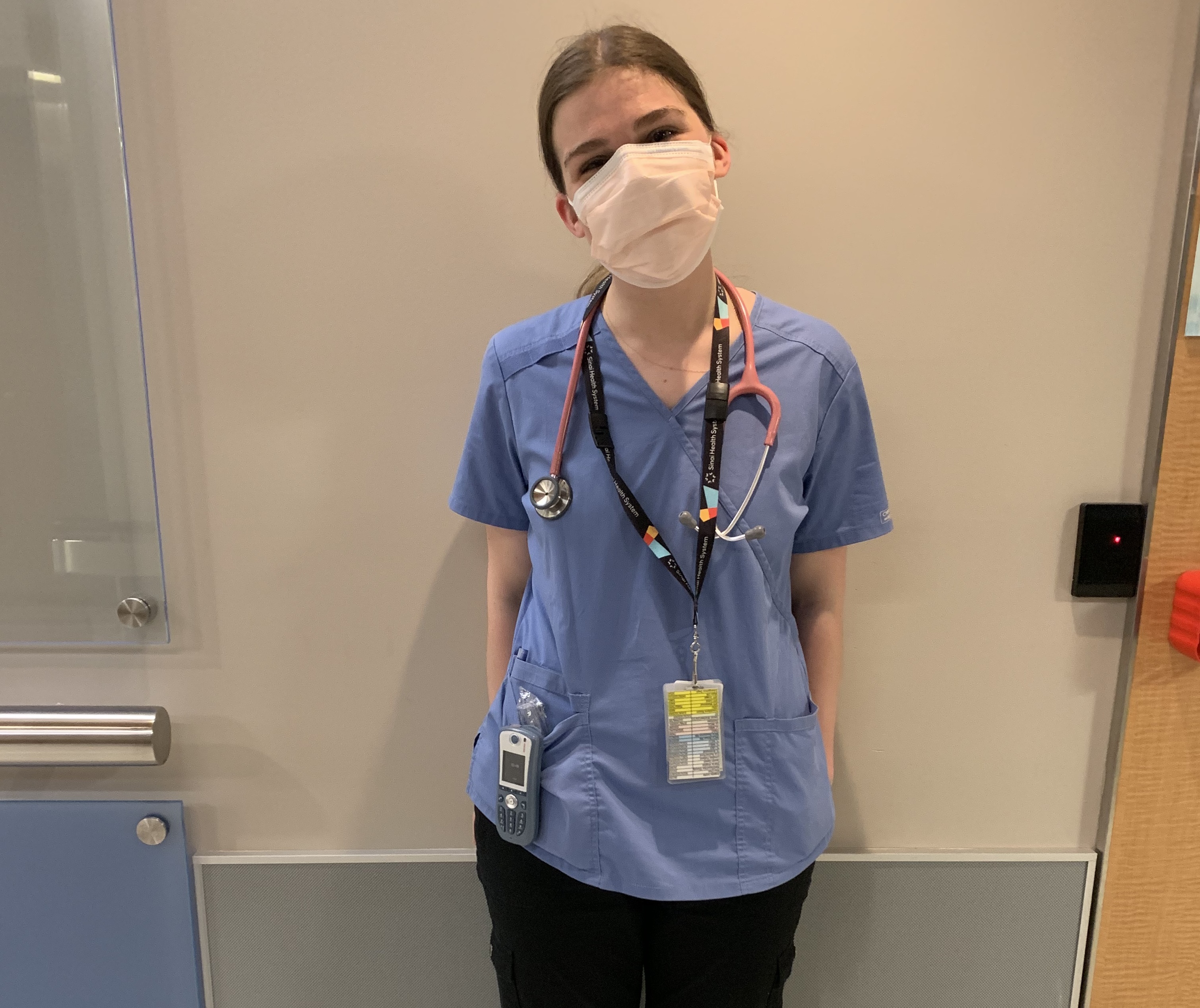 A nurse wearing scrubs and a mask standing in  a hallway, looking at the camera. She is also wearing a stethoscope, lanyard with employee ID badge and pager