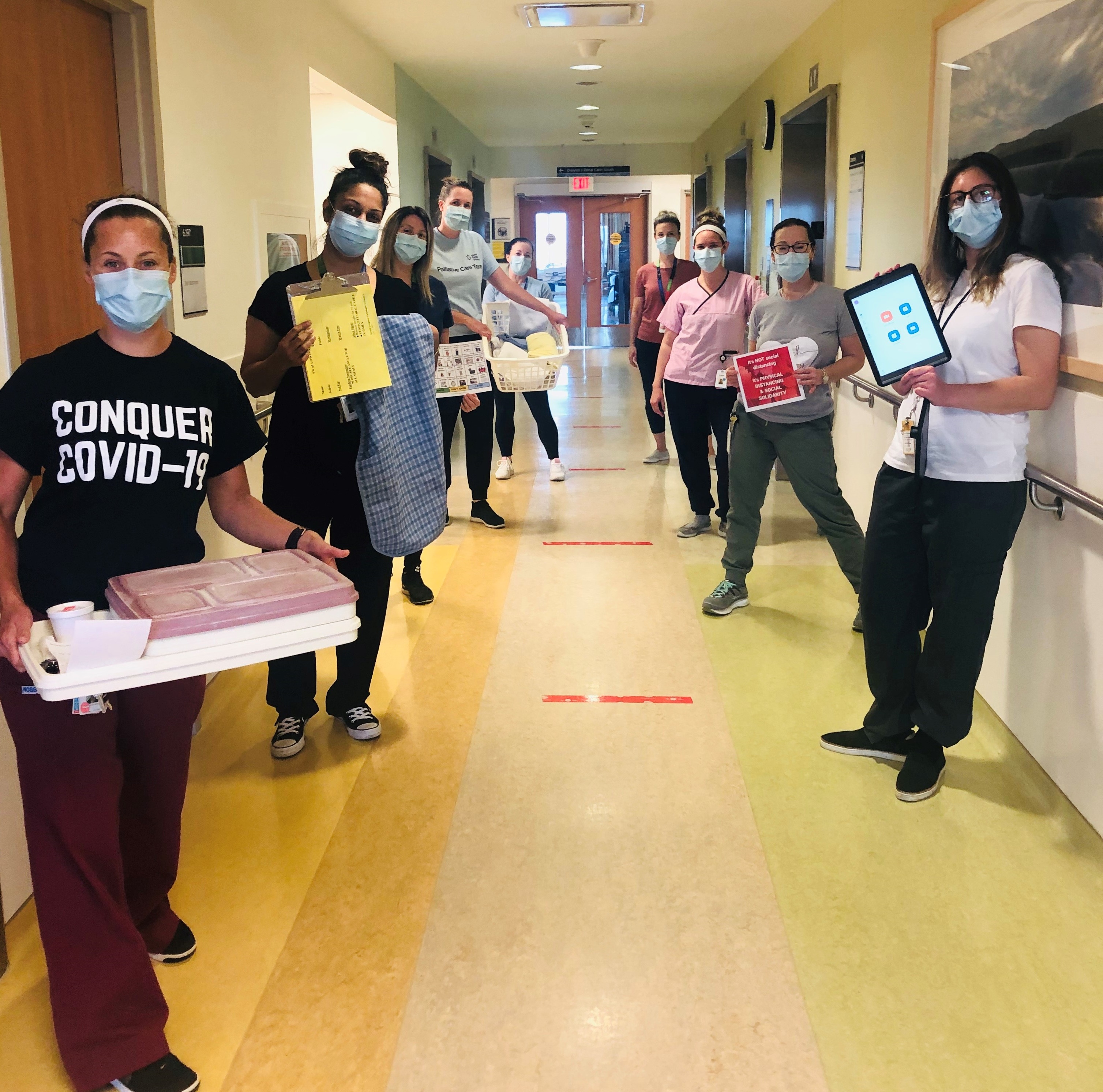 A group of hospital employees standing apart down either side of a hospital hallway. They are holding different objects such as meal trays, iPads laundry basket and looking at the camera.