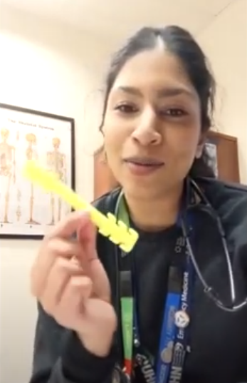A woman looking at the camera holding up a small yellow plastic strip that people in health care use to hold their masks on instead of putting the ear loops around their ears