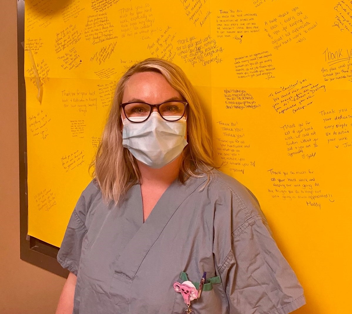 A health care worker wearing scrubs and a surgical mask stands in front of a large bright yellow poster on the wall. The poster has handwritten thank you messages on it