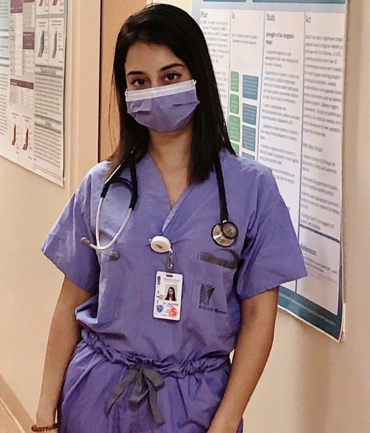 A physician wearing scrubs, a surgical mask and a stethoscope, standing in a hallway in a hospital. She is looking at the camera.