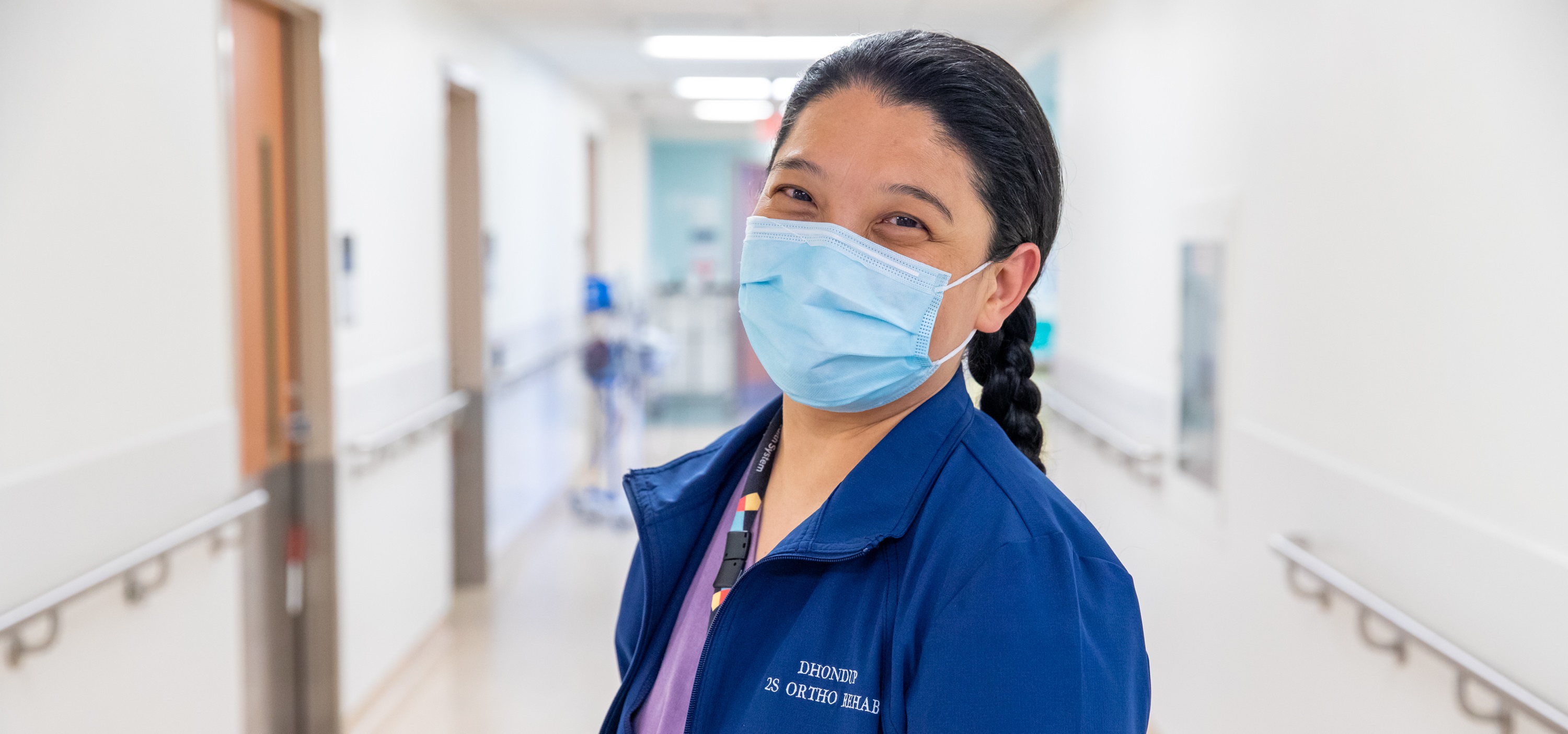 A nurse stands in a hospital hallway. She wearing a mask and looking at the camera, her eyes show that she is smiling under her mask.
