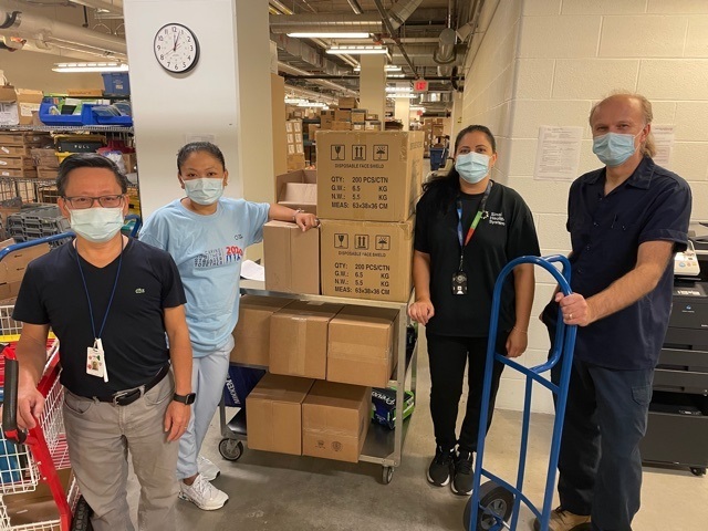 Four hospital employees in a shipping and receiving and storage area of a hospital. They are all facing the camera. They have mail carts and dollies next to them and cardboard boxes on shelves are visible in the background. 