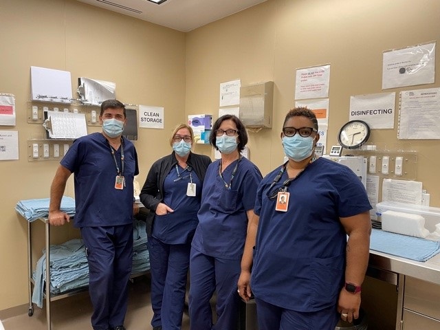 A group of people all wearing dark blue scrubs and surgical masks. They are in a hospital and there is hospital equipment behind them.
