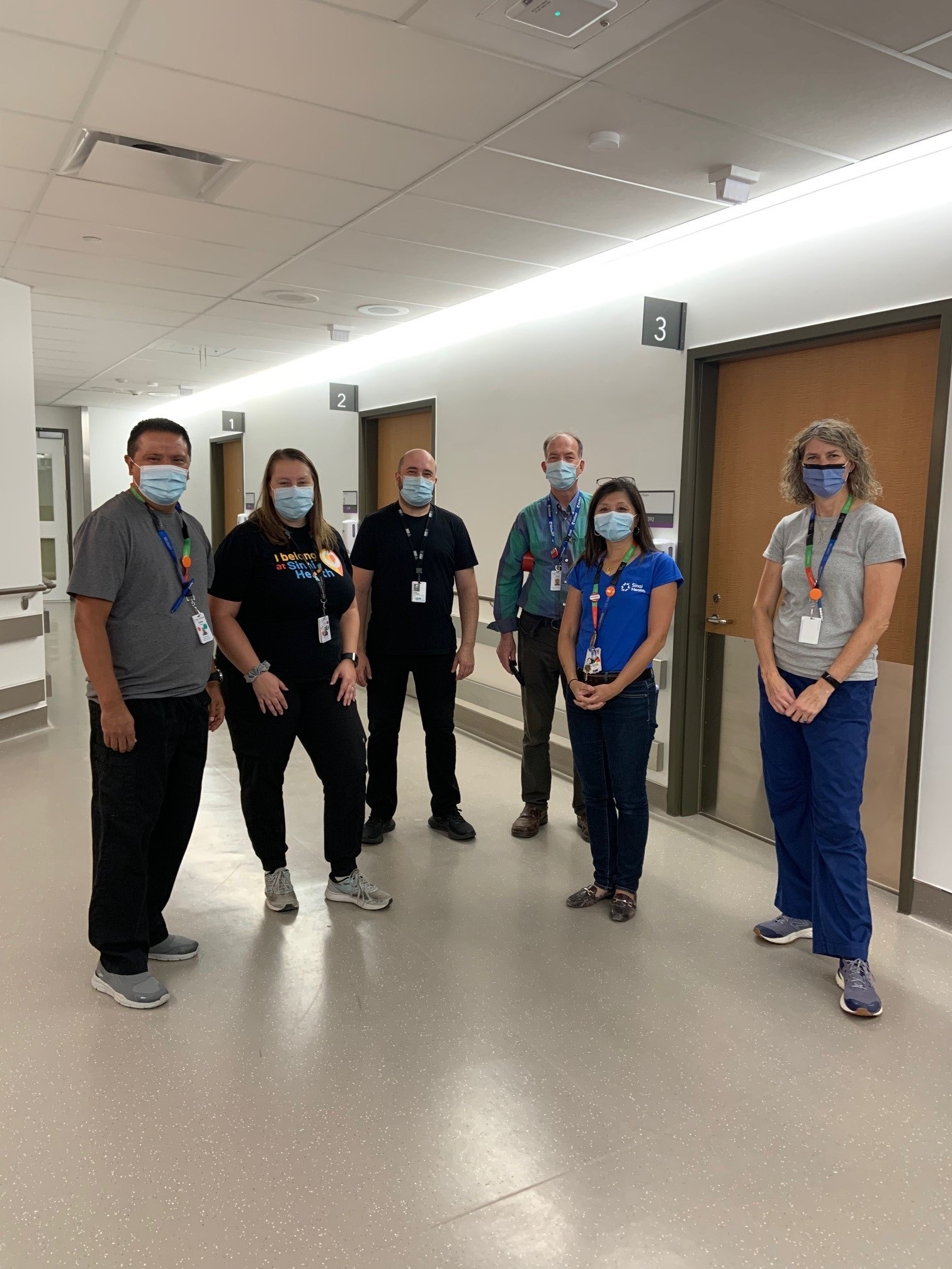 A group of people standing in a hall in a hospital. They are wearing masks and looking at the camera.