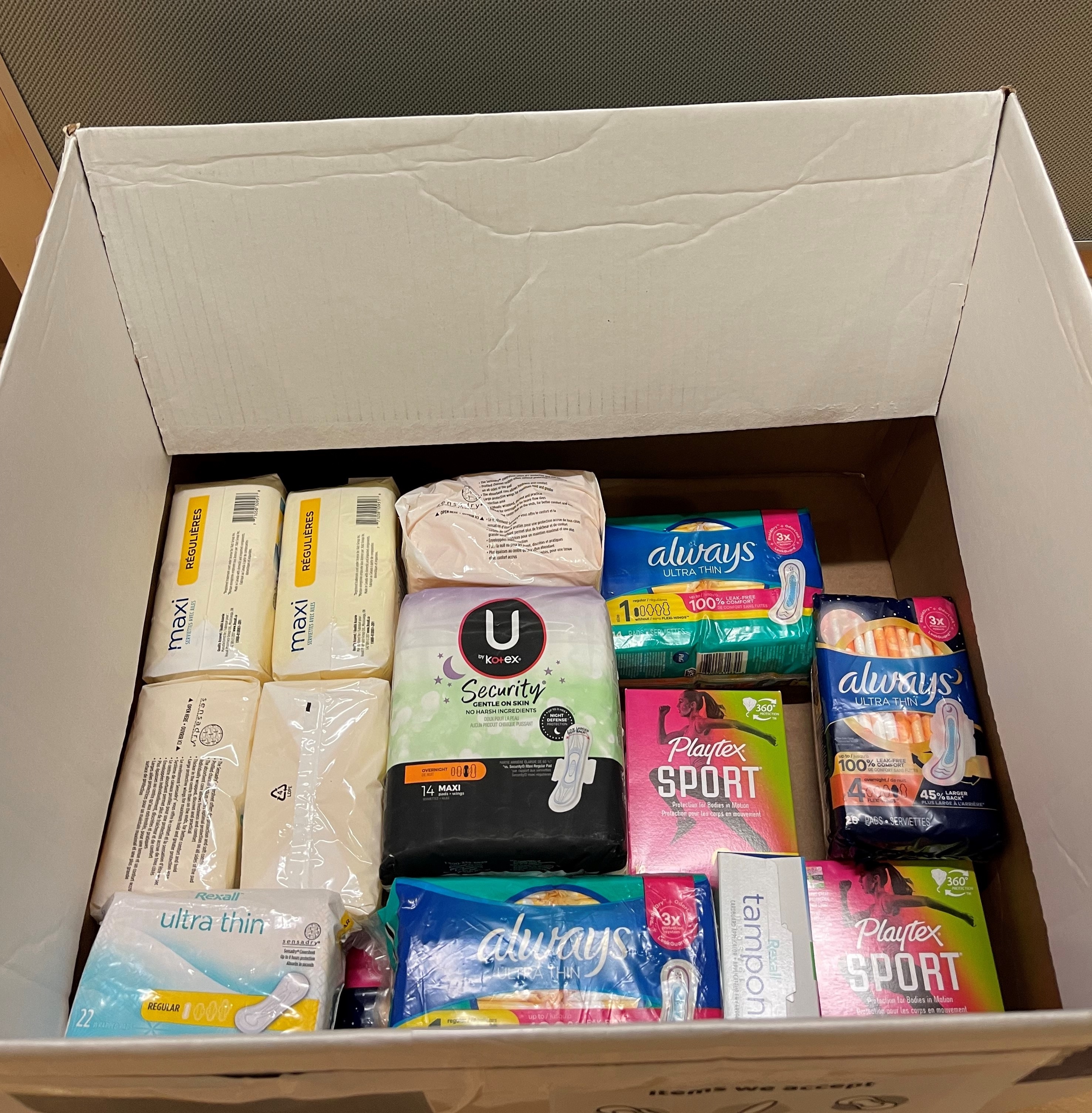 A cardboard box partially filled with packages of menstrual products of different types and brands 