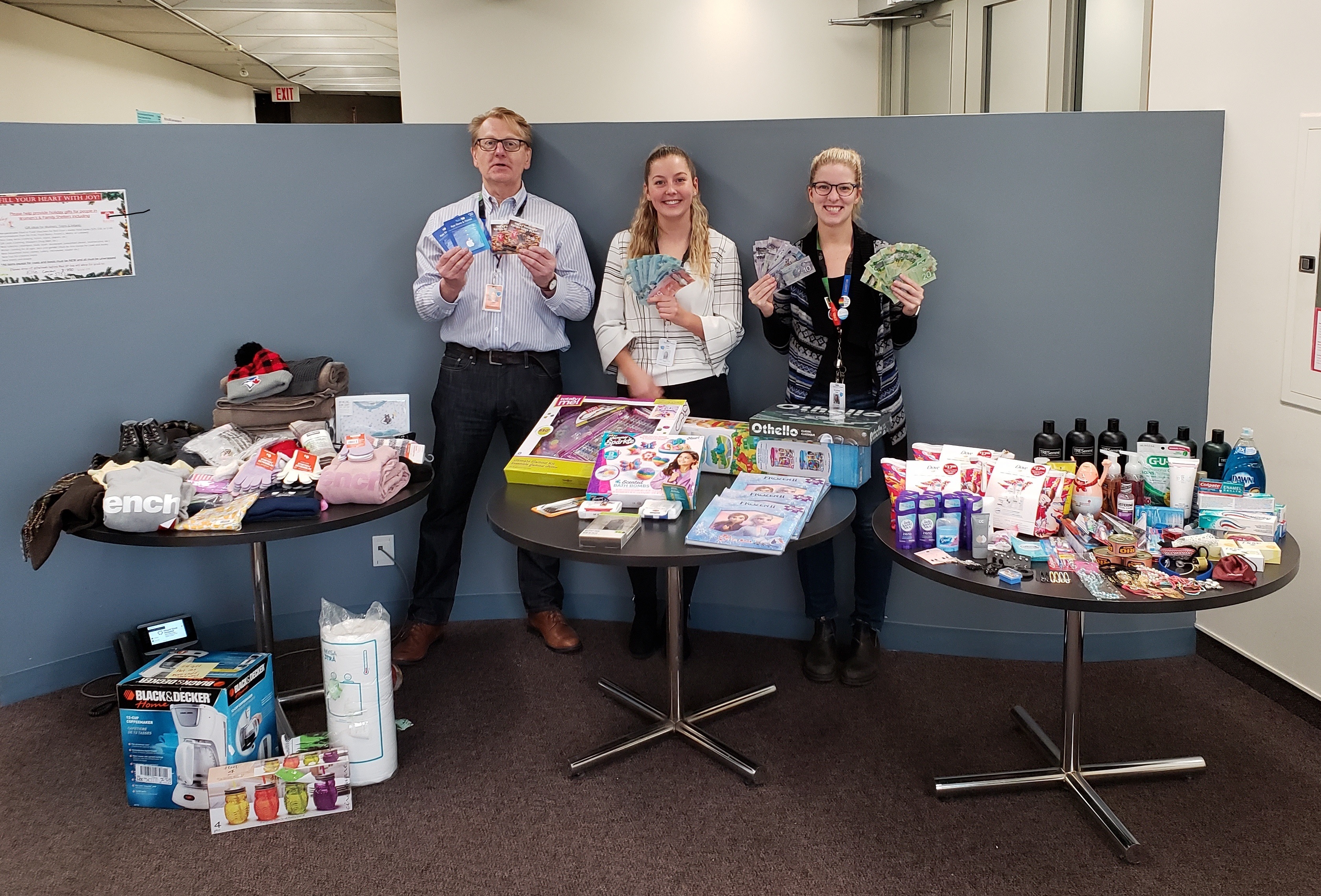 Three people in an office environment standing behind three round tables. The tables are filled with items, including scarves, hats, toys and personal care products donated to give to shelters during the holiday season. the people are also holding up gift cards and cash donations they have received.