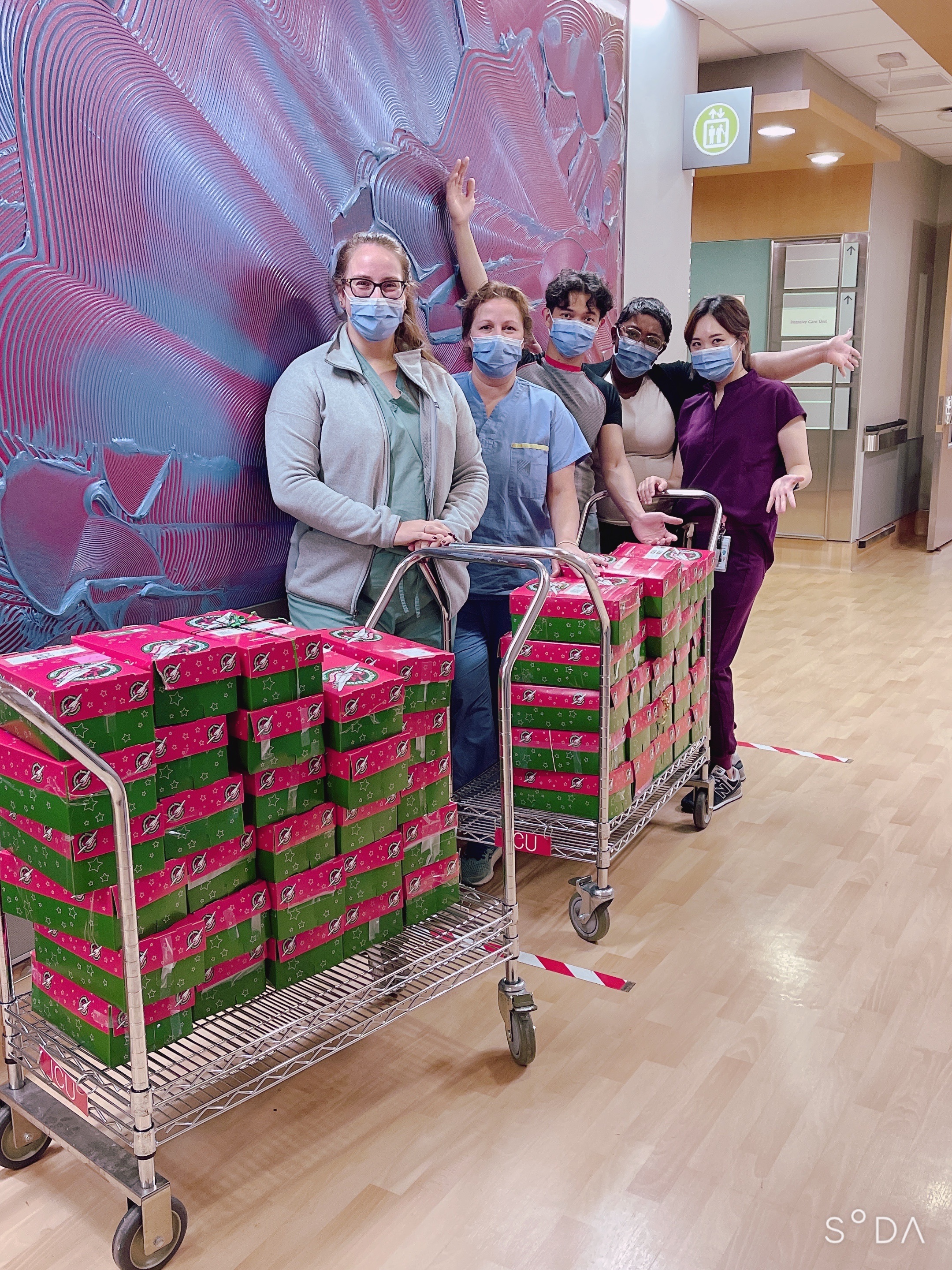 A group of five health care workers in a hallway in a hospital. they are standing next to two carts that each contain a large stack of gift wrapped shoe boxes neatly stacked. There are almost thirty shoe boxes on one cart