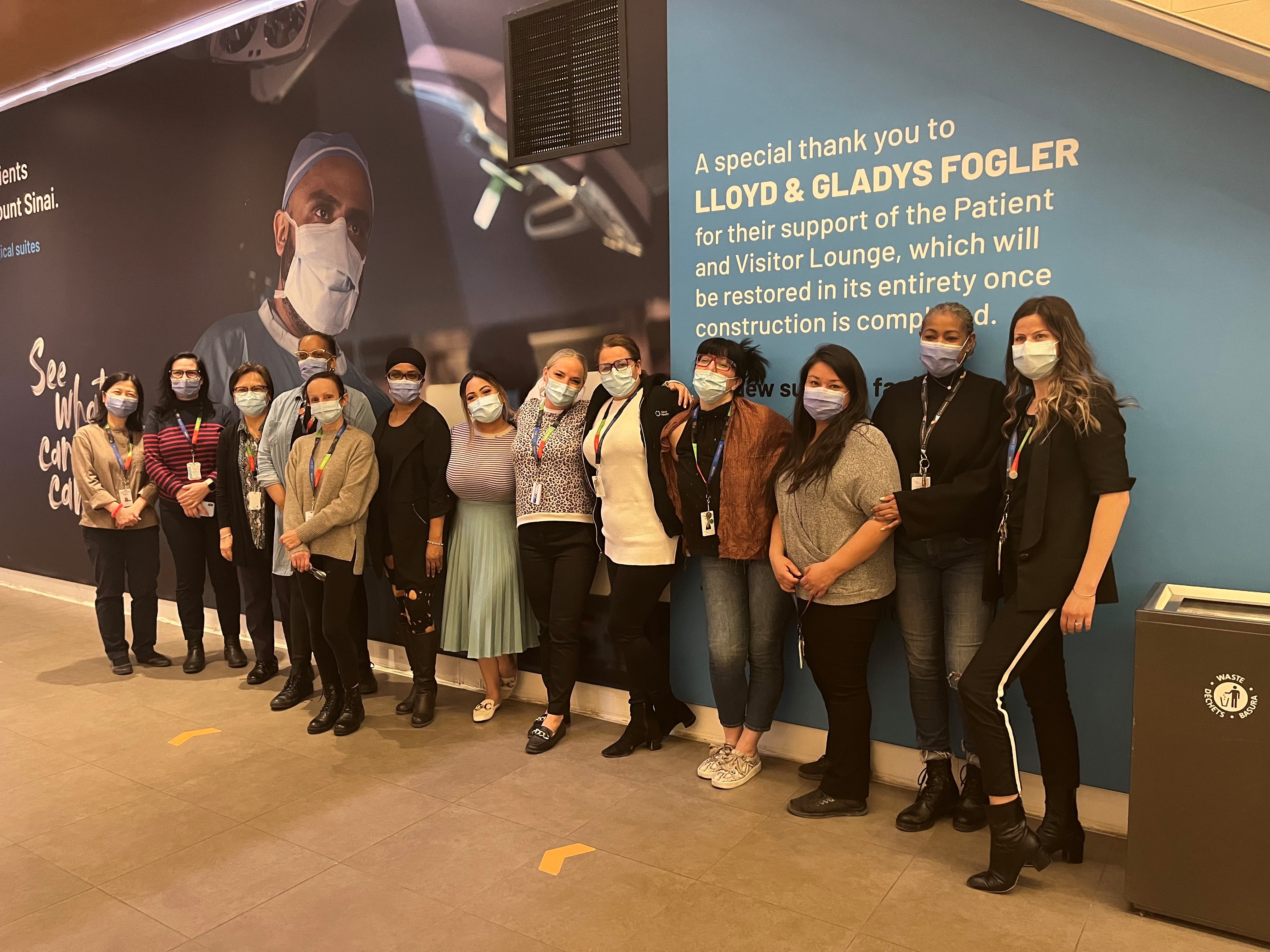 A group of health care professionals wearing medical procedure masks stand in a line along a wall in the corridor of a hospital. the wall is covered in a very large image showing a health care professional who is wearing a mask and scrubs like a surgeon wears.