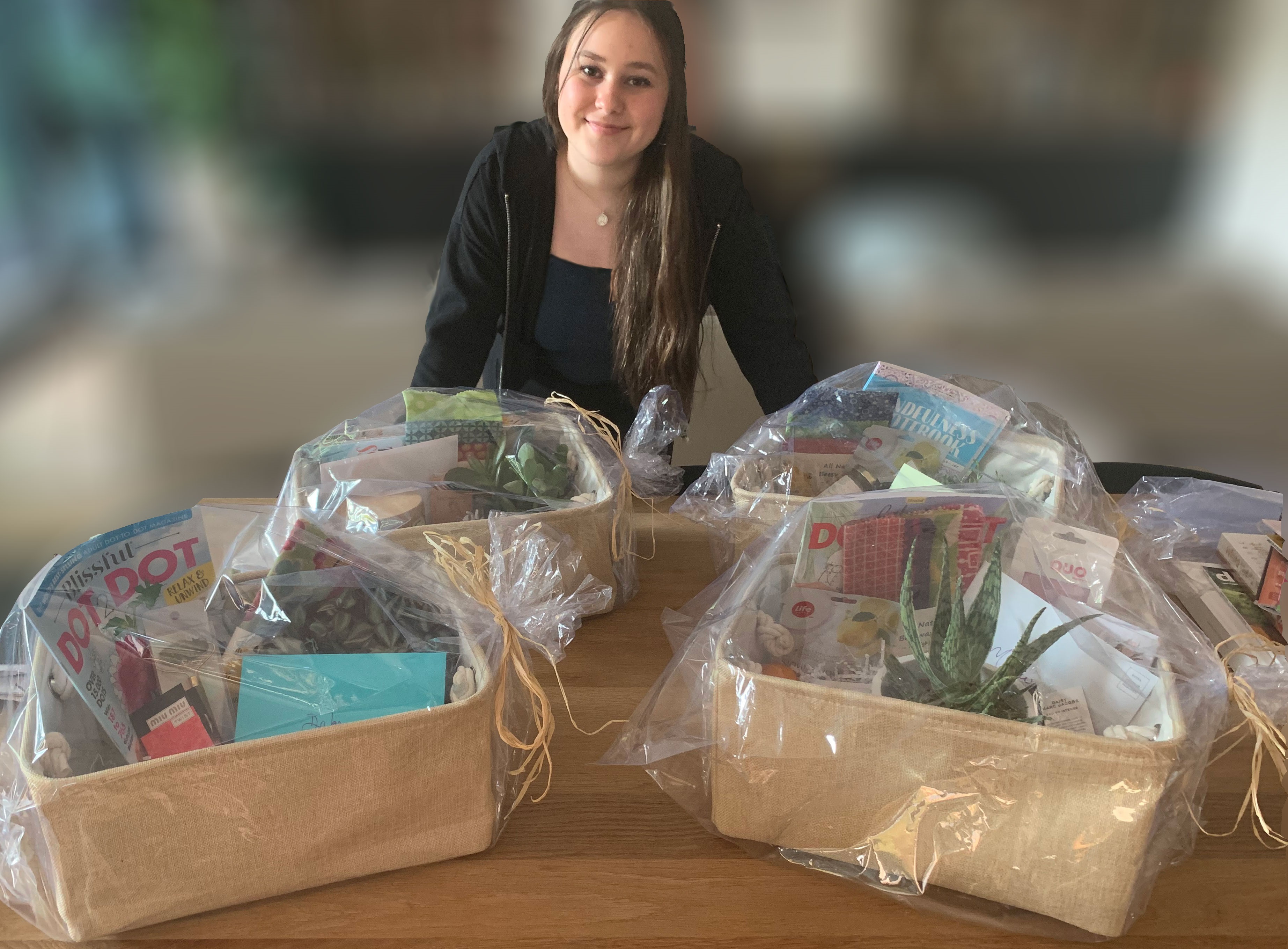 A young woman stands behind a table that is holding gift baskets wrapped in clear cellophane. The baskets contain items such as magazines, and small house plants. The woman is standing leaning on the table, facing the camera and smiling. The background is blurred.
