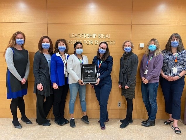 A group of health care professionals standing in a line looking at the camera. They are wearing medical procedure masks and two in the middle of the line are holding either side of a plaque they received for an award