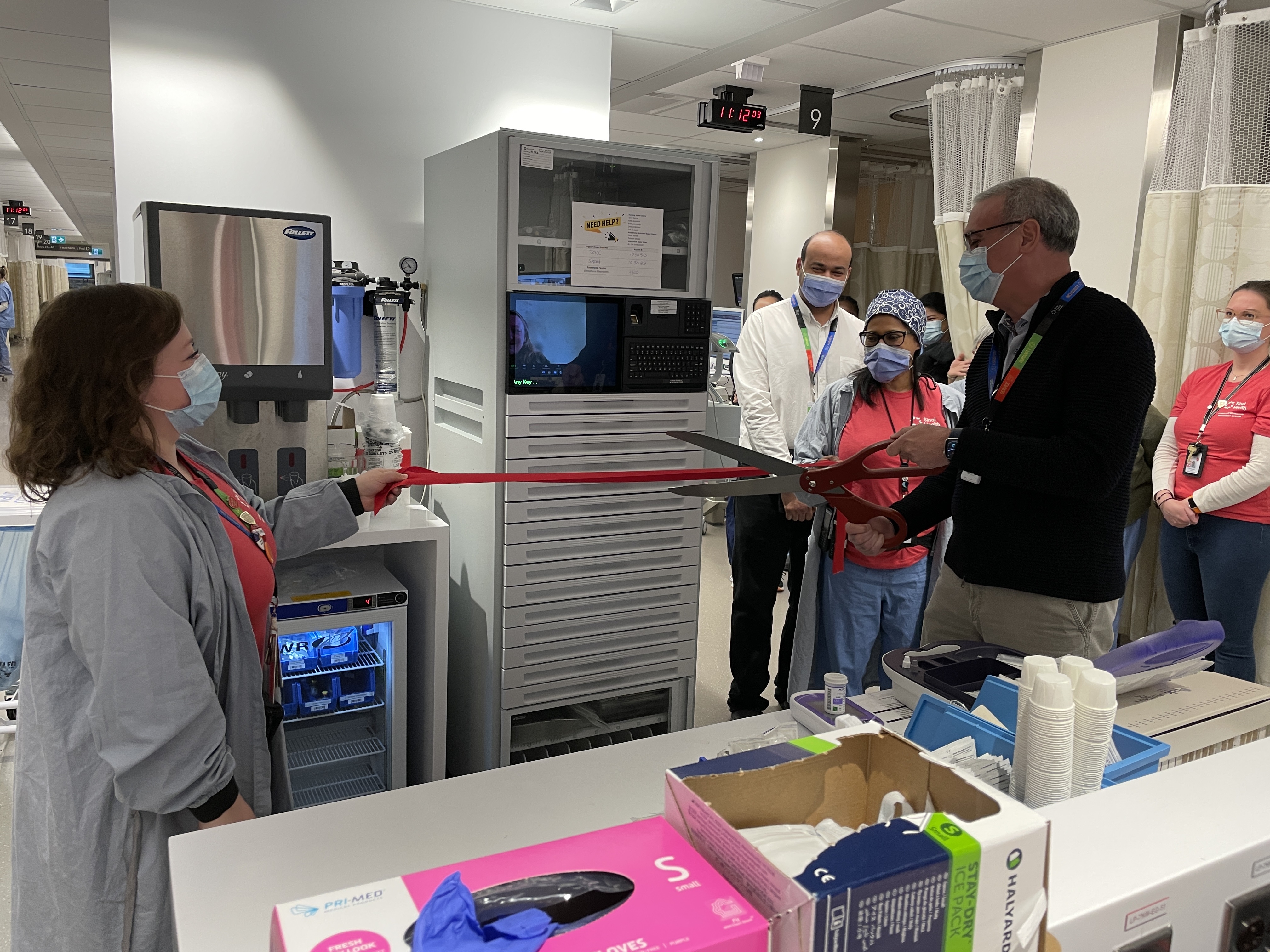 Dr. Gary Newton, president and CEO holds a large pair of scissors poised to cut through a red ribbon held in front of an automated medication dispensing machine. The machine is a cabinet with drawers and is similar in size to a refrigerator . Two women are standing on either side of the machine holding up each end of the ribbon. Other employees look on in the background
