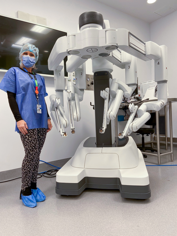 A female health care worker wearing surgical scrubs including cap and surgical mask stands next to a large surgical robot The machine is taller than the woman and has four large mechanical arms connected to a central post