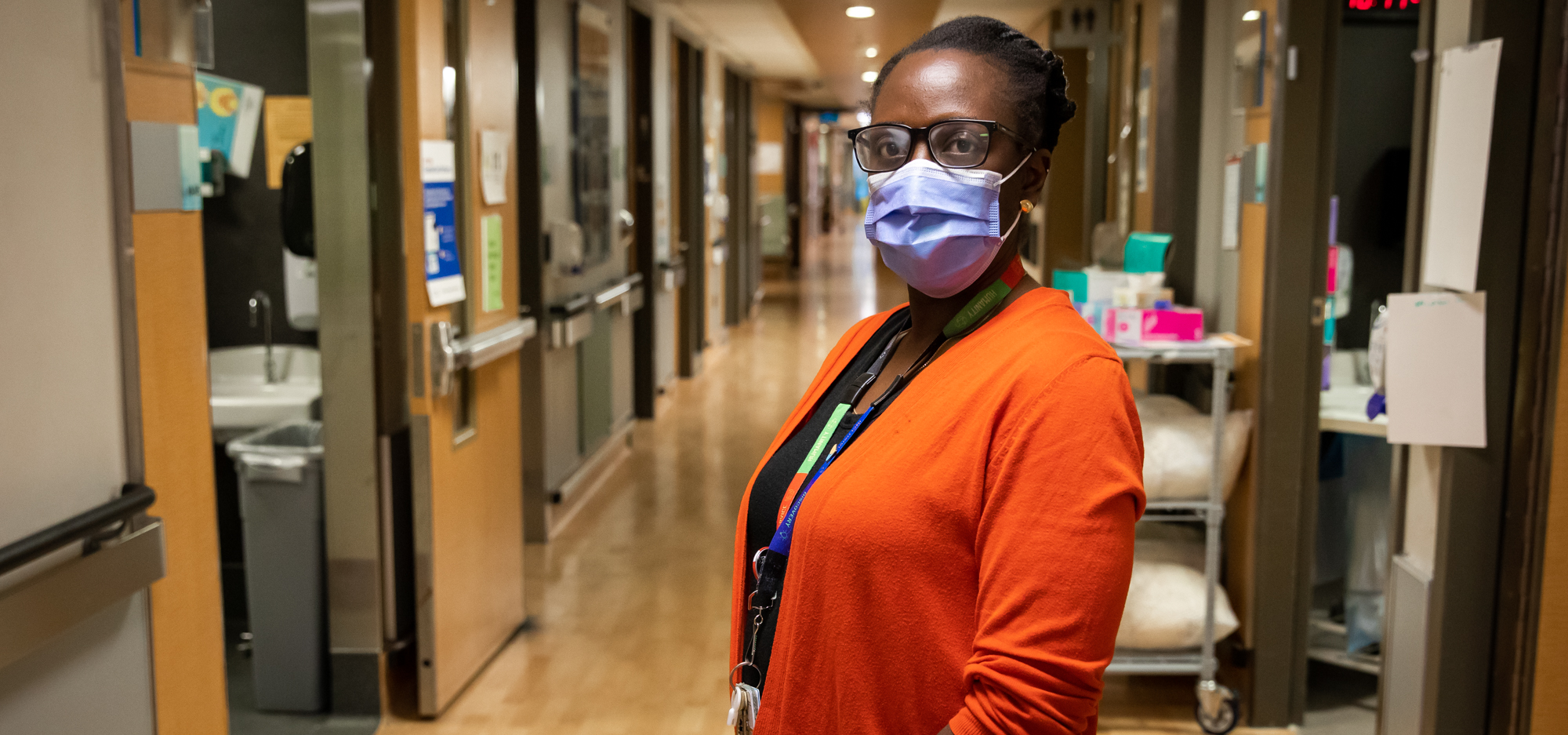Nana Asomaning, a patient care manager at Mount Sinai Hospital, stands in the hall way of the unit where she works. She is looking at the camera and wearing a medical grade procedure maskl procedure mask.