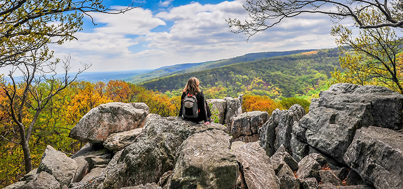 A woman wearing a backpack is sitting on a rocky overlook with her back to the camera. She is overlooking the Appalachian Mountains covered in trees, which are changing to fall colours.  