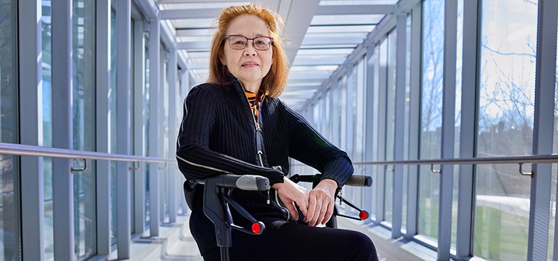 A woman sits on a rollator mobility device. She is surrounded on both sides by the windows of an indoor walkway. She is looking at the camera smiling with her hands clasped together. She has auburn hair, glasses and is wearing a black.