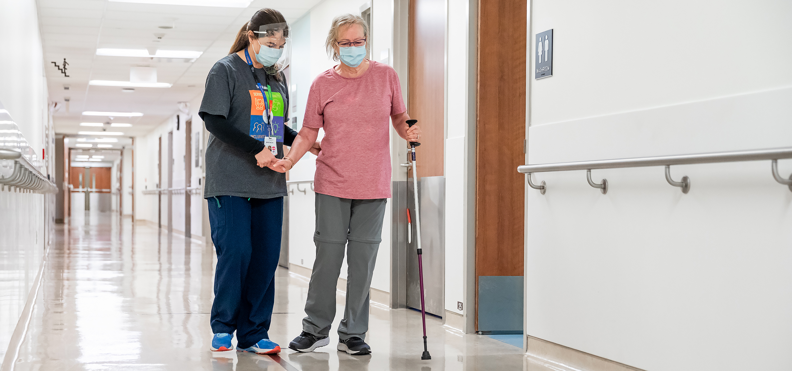 A health-care provider and a patient are walking down the hallway of a hospital. The patient is an older adult woman and she is walking towards the camera with a walking pole in her left hand. The health care provider walks on the patient's right side, holding her hand and offering support