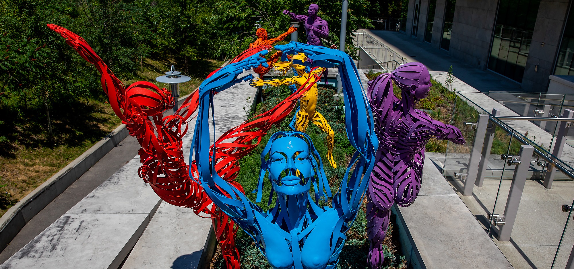 a group of six metal sculptures in the Max Tannebaum Sculpture garden. They are human figures made out of narrow strips of metal in bright colours. Each figure is depicted as if it is frozen in the middle of motion. The sculpture closest to the camera is dancing